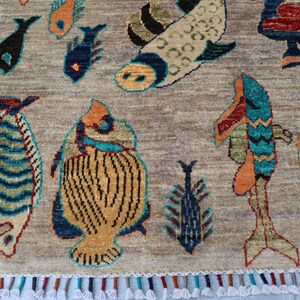 May include: A rectangular area rug with a light brown background featuring various stylised fish motifs in shades of blue, orange, yellow, and green. The rug is finished with a decorative fringe along the bottom edge containing multicoloured tassels.