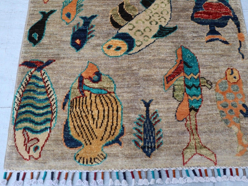 May include: A rectangular area rug with a light brown background featuring various stylised fish motifs in shades of blue, orange, yellow, and green. The rug is finished with a decorative fringe along the bottom edge containing multicoloured tassels.