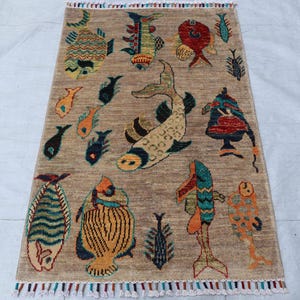 May include: A rectangular area rug with a tan background featuring a variety of colourful fish illustrations in shades of blue, orange, red, and yellow. The rug has a textured surface and is finished with short, multi-coloured fringe at both ends.