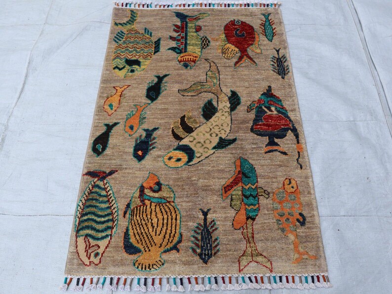 May include: A rectangular area rug with a tan background featuring a variety of colourful fish illustrations in shades of blue, orange, red, and yellow. The rug has a textured surface and is finished with short, multi-coloured fringe at both ends.