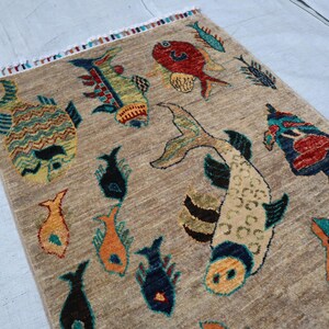 May include: A rectangular area rug with a tan, textured background featuring a variety of colourful fish illustrations in shades of red, blue, orange, and green. The rug has a short fringe along the top edge with multi-coloured thread accents.