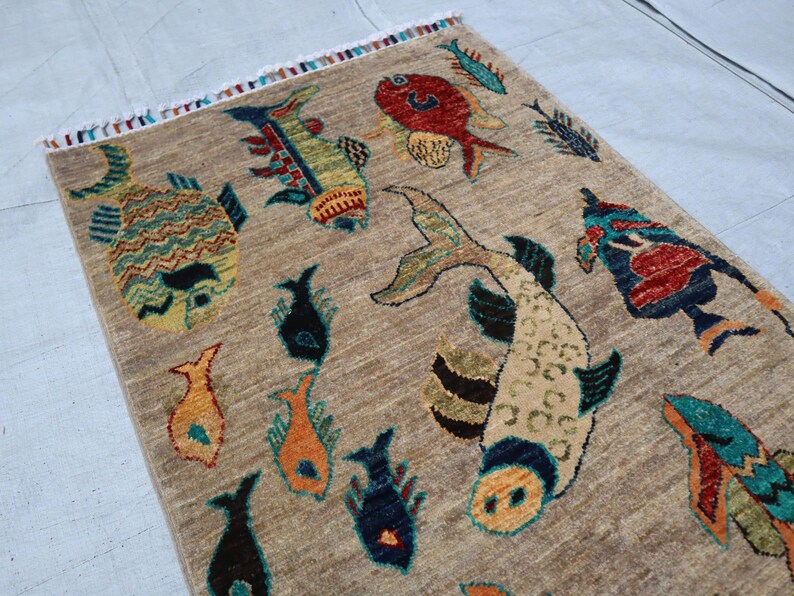 May include: A rectangular area rug with a tan, textured background featuring a variety of colourful fish illustrations in shades of red, blue, orange, and green. The rug has a short fringe along the top edge with multi-coloured thread accents.