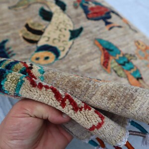 May include: A beige area rug featuring a repeating pattern of stylised fish in shades of blue, orange, and black. The rug has a thick, tufted pile texture and includes decorative fringe along the edge with multi-coloured threads in red and blue.