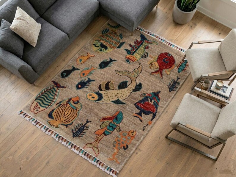 May include: A rectangular area rug featuring a tan background with a variety of colourful fish illustrations in shades of red, blue, yellow, and green. The rug has a textured surface and is finished with decorative fringe along the shorter edges.