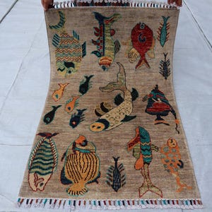 May include: A rectangular area rug with a light brown background featuring a variety of colourful fish illustrations in shades of red, blue, yellow, and green. The rug has a fringed edge with multi-coloured tassels along the bottom border.