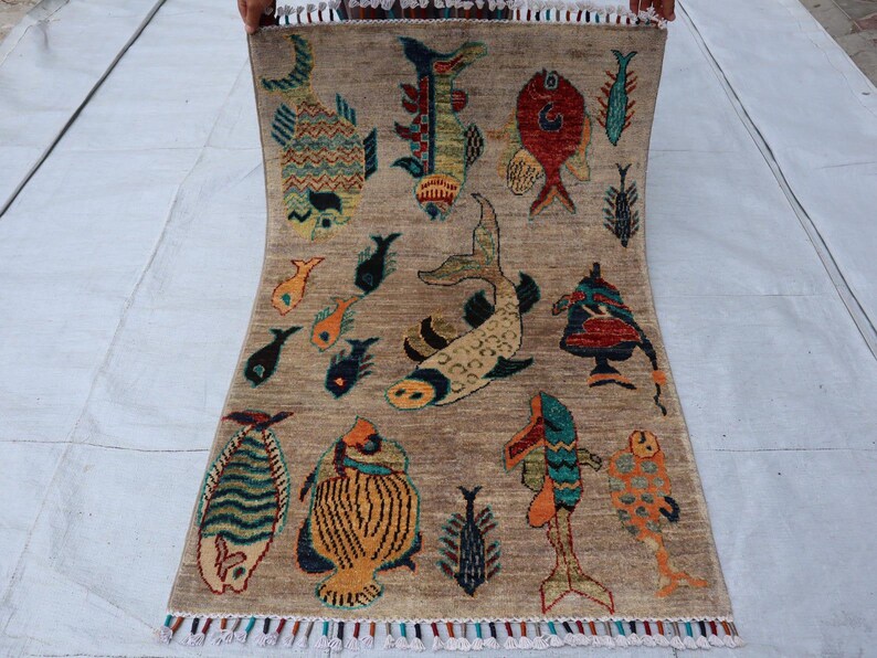 May include: A rectangular area rug with a light brown background featuring a variety of colourful fish illustrations in shades of red, blue, yellow, and green. The rug has a fringed edge with multi-coloured tassels along the bottom border.