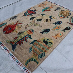 May include: A rectangular area rug with a beige background featuring a pattern of various fish in colours including red, yellow, blue, and green. The rug has a textured surface and is finished with short, multi-coloured fringe along the two shorter ends.