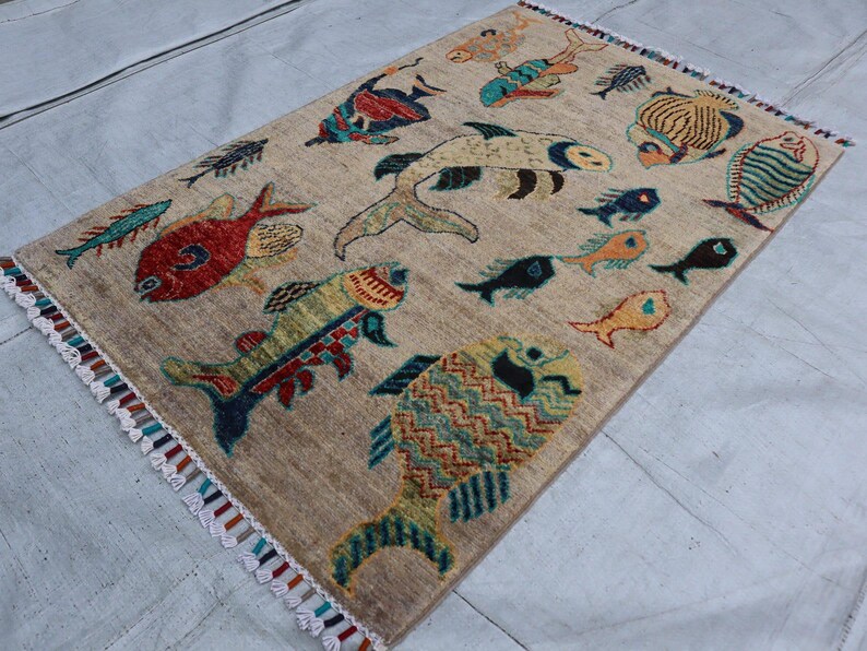 May include: A rectangular area rug with a beige background featuring a pattern of various fish in colours including red, yellow, blue, and green. The rug has a textured surface and is finished with short, multi-coloured fringe along the two shorter ends.