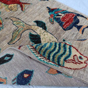 May include: A beige area rug featuring a repeating pattern of stylised fish in vibrant colours including red, blue, green, and orange. The rug has a folded corner revealing a textured surface and a decorative fringe edge with multi-coloured tassels.