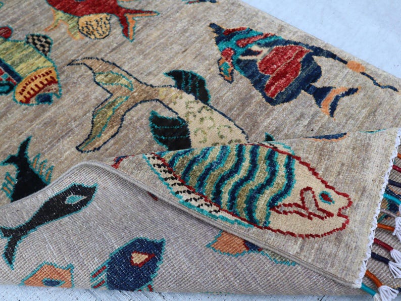 May include: A beige area rug featuring a repeating pattern of stylised fish in vibrant colours including red, blue, green, and orange. The rug has a folded corner revealing a textured surface and a decorative fringe edge with multi-coloured tassels.