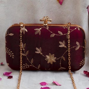 May include: A rectangular burgundy clutch with gold floral embroidery and a gold chain strap. The clutch has a gold clasp and is decorated with gold leaves and flowers. The background has scattered pink flower petals.