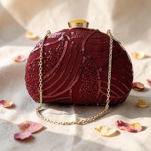 Puede incluir: Un bolso de mano color burdeos con intrincados bordados de lentejuelas en patrones ondulados. El bolso cuenta con un marco de metal dorado, una correa de cadena dorada a juego y un cierre circular dorado con una pieza superior amarilla.