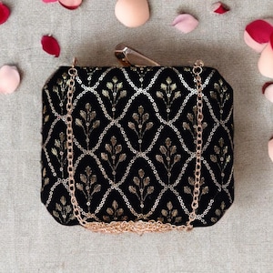 Op de afbeelding: Zwarte rechthoekige clutch met een gouden bladpatroon en een gouden ketting. De clutch heeft een zilveren sluiting en is versierd met gouden pailletten. De achtergrond bevat roze bloemblaadjes en ronde, lichtroze objecten.