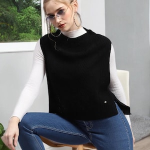 Könnte beinhalten: Eine schwarze ärmellose Weste mit hohem Ausschnitt. Die Weste wird über einem weißen Rollkragenpullover getragen und mit einer blauen Jeans kombiniert. Das Model trägt eine Sonnenbrille und Creolen. Das Outfit ist lässig und stilvoll.