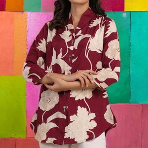 Può includere: Camicia bordeaux con bottoni e motivo floreale color crema. La camicia a maniche lunghe ha un colletto a revers ed è abbottonata. La modella indossa pantaloni bianchi e orecchini di perle. Lo sfondo è un colorato patchwork di quadrati dipinti.