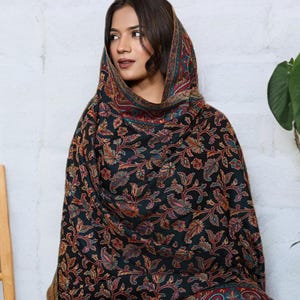 May include: A black shawl featuring a detailed floral design in red, blue, and green. The shawl is worn over the head and shoulders, with a patterned border. The material looks soft and drapes well.
