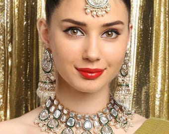 May include: A woman wearing a gold-toned necklace, earrings, and headpiece. The jewelry features clear stones and pearls. She is wearing a green sequined wrap. The background is a gold curtain.