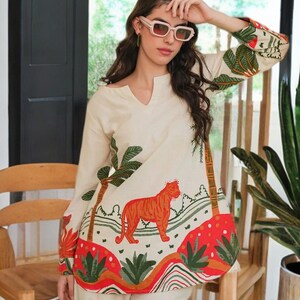 May include: Cream-colored long-sleeved top and pants set. The top features a V-neck and a vibrant tiger illustration with palm trees and tropical foliage in orange, red, and green. The pants are a matching cream color.
