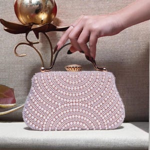 May include: A blush-colored beaded clutch with a decorative handle. The clutch is covered in small, round, white beads arranged in a fan pattern. The handle is gold-toned, and the clasp is a small, round, gold-toned jewel.