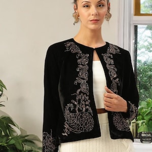 Può includere: Giacca in velluto nero con ricami color rame intricati. La giacca aperta presenta motivi dettagliati sul davanti e sulle maniche. Indossata sopra un top bianco e una gonna color crema, creando un contrasto di trame e colori.
