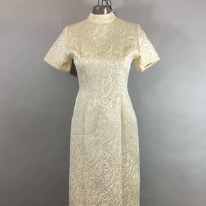 May include: A vintage ivory-colored dress with a high neckline and short sleeves. The dress features an all-over embossed floral pattern. The dress is floor-length and appears to be made of a textured fabric, likely suitable for formal occasions.