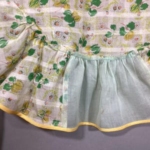 May include: A vintage apron with a floral print and a light blue ruffle. The apron has a yellow trim and a pocket.