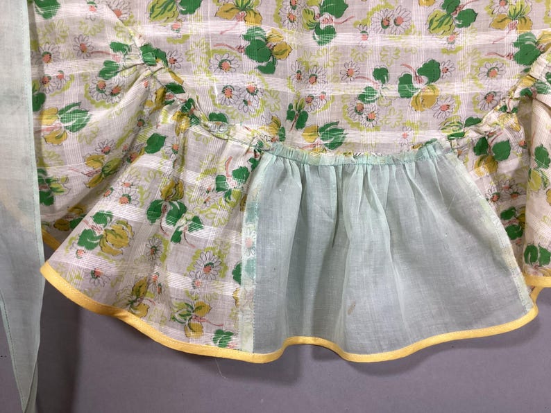 May include: A vintage apron with a floral print and a light blue ruffle. The apron has a yellow trim and a pocket.