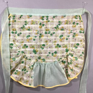 May include: A vintage green and yellow floral print apron with a ruffled bottom and yellow trim. The apron has two long ties that are tied at the waist.