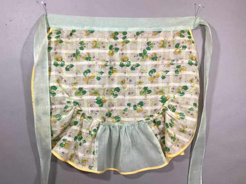 May include: A vintage green and yellow floral print apron with a ruffled bottom and yellow trim. The apron has two long ties that are tied at the waist.