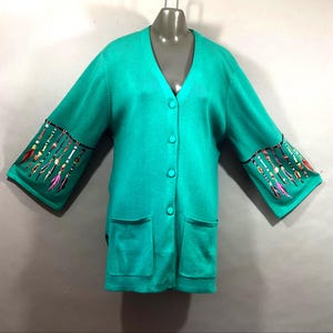 May include: Teal cardigan with a V-neck, button closure, and two front pockets. The sleeves feature a decorative design with colorful feathers and beads. The cardigan is displayed on a mannequin.