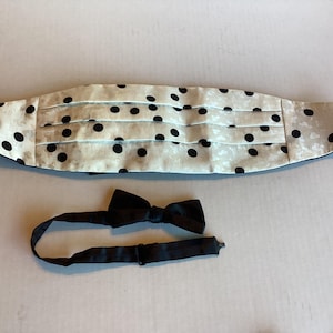 May include: A cummerbund featuring a cream-colored fabric with a subtle textured pattern and black polka dots, accompanied by a matching solid black bow tie with an adjustable strap and a metal sliding buckle for secure fastening.