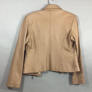 May include: A tan leather jacket with a classic design. The jacket features a collared neckline, long sleeves, and a tailored fit. The back of the jacket is visible, showcasing the seams and construction details. The jacket is hanging on a metal hanger.