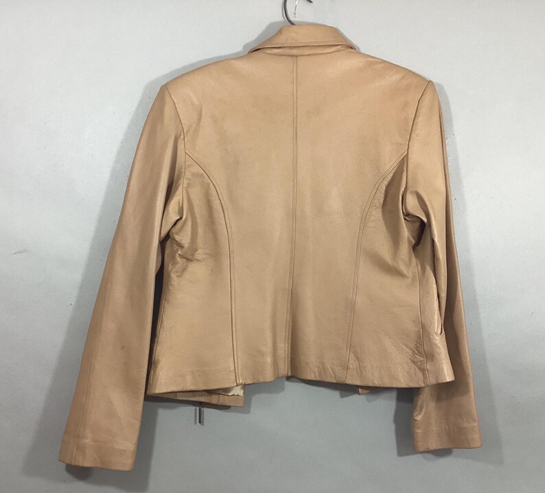 May include: A tan leather jacket with a classic design. The jacket features a collared neckline, long sleeves, and a tailored fit. The back of the jacket is visible, showcasing the seams and construction details. The jacket is hanging on a metal hanger.