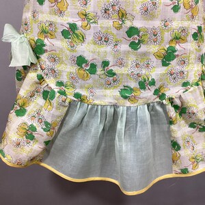 May include: A vintage apron with a white background and a floral pattern of green leaves and yellow flowers. The apron has a ruffled bottom and yellow trim. The apron has two green fabric ties.