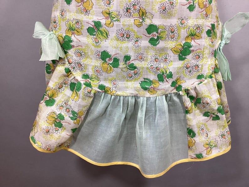 May include: A vintage apron with a white background and a floral pattern of green leaves and yellow flowers. The apron has a ruffled bottom and yellow trim. The apron has two green fabric ties.