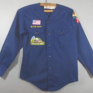 May include: A navy blue Cub Scout shirt with long sleeves, a button-down front, and two chest pockets. The shirt features an American flag patch, the words "CUB SCOUT", and other patches. A vintage item.