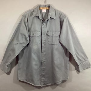 May include: A light gray button-down shirt with long sleeves, two chest pockets, and a collar. The shirt is hanging on a hanger. The label reads "RAY DYERS & CLE".