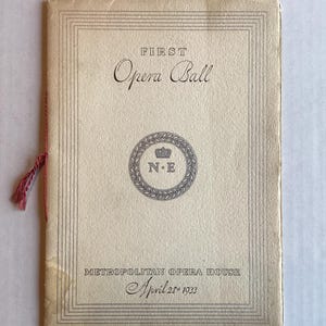May include: A cream-colored paper program for the First Opera Ball at the Metropolitan Opera House on April 28, 1933. The cover features black text, a central circular emblem with a crown and the letters N.E, and a red string binding on the left.