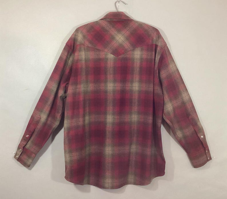 Puede incluir: Camisa de manga larga con un estampado a cuadros en rojo, beige y marr&oacute;n. La camisa est&aacute; abotonada y colgada de una percha met&aacute;lica. La parte trasera de la camisa es visible, mostrando la pieza del canes&uacute; y los pu&ntilde;os abotonados.