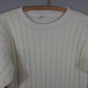 May include: A white quilted sweater with a ribbed texture. The sweater has a crew neck and long sleeves.