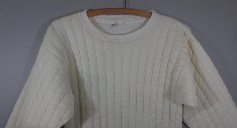 May include: A white quilted sweater with a ribbed texture. The sweater has a crew neck and long sleeves.