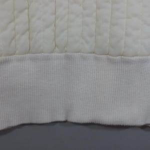 May include: Close-up of the bottom edge of a white quilted garment. The edge is finished with a white ribbed knit band.