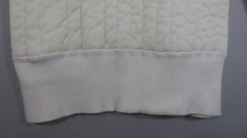 May include: Close-up of the bottom edge of a white quilted garment. The edge is finished with a white ribbed knit band.