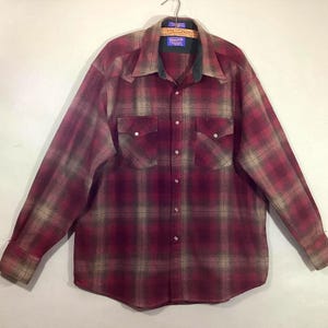 May include: A long-sleeved, button-down plaid shirt in shades of burgundy, tan, and green. The shirt has two chest pockets and a collar with a dark green underside. The shirt is hanging on a wooden hanger.