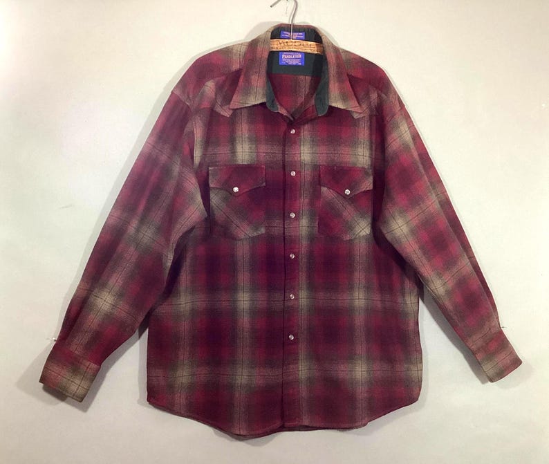 Puede incluir: Una camisa de franela a cuadros con botones y mangas largas en tonos burdeos, beige y verde. La camisa tiene dos bolsillos en el pecho y un cuello con la parte inferior verde oscuro. La camisa est&aacute; colgada en una percha de madera.