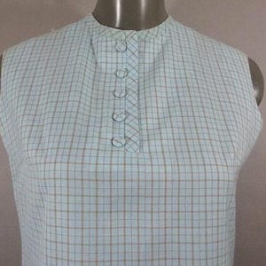 May include: A sleeveless blouse with a checkered pattern in shades of blue and brown. The blouse has a high, round neckline and a button closure at the front.