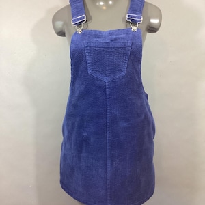 May include: A blue corduroy overall dress with adjustable straps and a front pocket. The dress is displayed on a mannequin and features silver-colored hardware on the straps.
