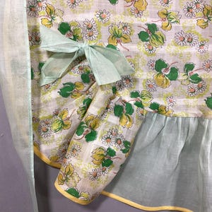 May include: A vintage apron with a green and white floral print. The apron has a light green ruffle at the bottom and a light green bow at the waist.
