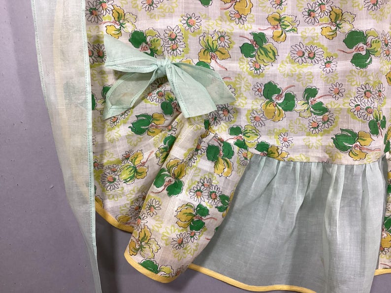 May include: A vintage apron with a green and white floral print. The apron has a light green ruffle at the bottom and a light green bow at the waist.