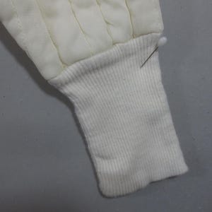 May include: Close-up of a white quilted jacket sleeve with a ribbed knit cuff. The cuff is folded back and secured with a pin.